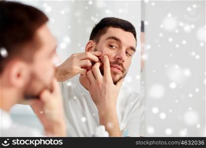 beauty, hygiene, skin problem and people concept - young man looking to mirror and squeezing pimple at home bathroom over snow