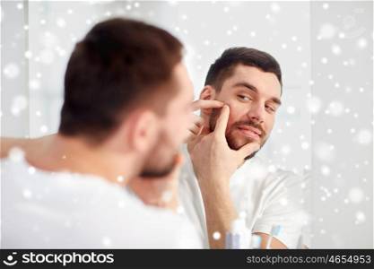 beauty, hygiene, skin problem and people concept - young man looking to mirror and squeezing pimple at home bathroom over snow