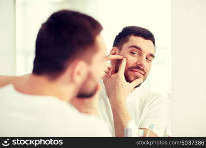 beauty, hygiene, skin problem and people concept - young man looking to mirror and squeezing pimple at home bathroom. man squeezing pimple at bathroom mirror