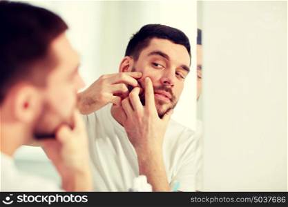 beauty, hygiene, skin problem and people concept - young man looking to mirror and squeezing pimple at home bathroom. man squeezing pimple at bathroom mirror