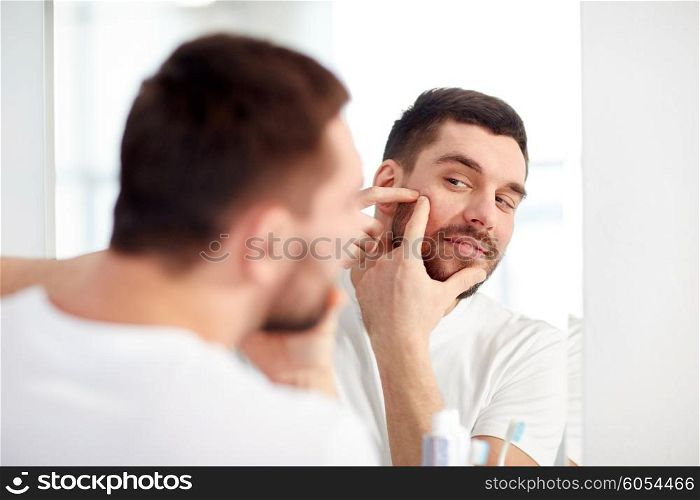 beauty, hygiene, skin problem and people concept - young man looking to mirror and squeezing pimple at home bathroom