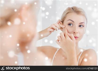 beauty, hygiene, skin problem and people concept - young woman looking to mirror and squeezing pimple at home bathroom over snow