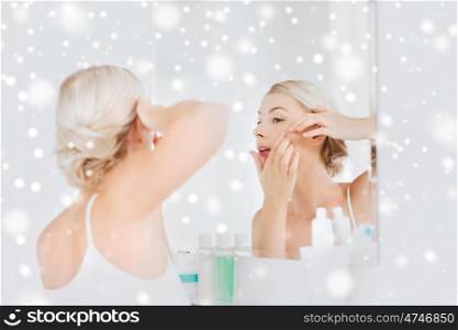 beauty, hygiene, skin problem and people concept - young woman looking to mirror and squeezing pimple at home bathroom over snow