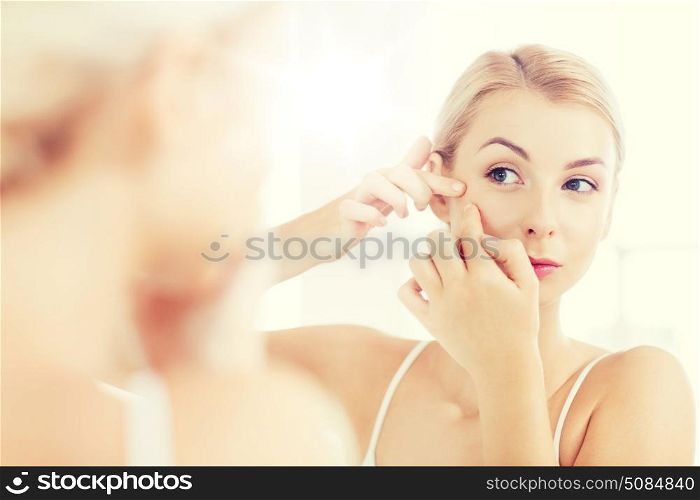 beauty, hygiene, skin problem and people concept - young woman looking to mirror and squeezing pimple at home bathroom. woman squeezing pimple at bathroom mirror. woman squeezing pimple at bathroom mirror