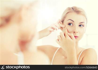 beauty, hygiene, skin problem and people concept - young woman looking to mirror and squeezing pimple at home bathroom. woman squeezing pimple at bathroom mirror. woman squeezing pimple at bathroom mirror