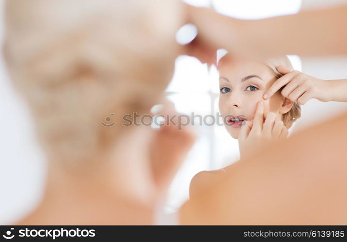 beauty, hygiene, skin problem and people concept - young woman looking to mirror and squeezing pimple at home bathroom