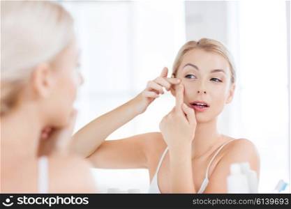 beauty, hygiene, skin problem and people concept - young woman looking to mirror and squeezing pimple at home bathroom