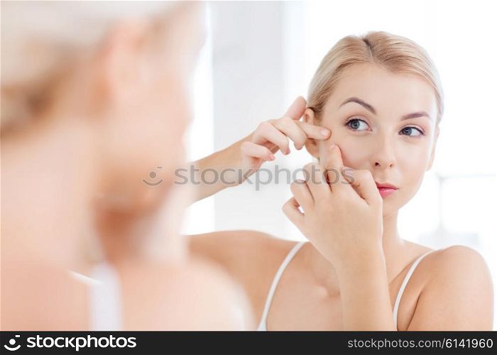 beauty, hygiene, skin problem and people concept - young woman looking to mirror and squeezing pimple at home bathroom