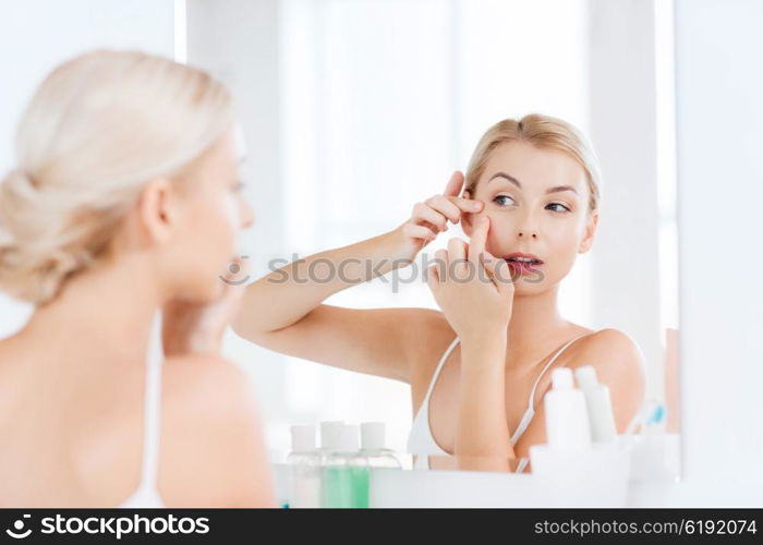 beauty, hygiene, skin problem and people concept - young woman looking to mirror and squeezing pimple at home bathroom