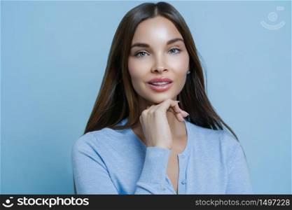 Beauty is in natural look. Portrait of pleasant looking European woman keeps hand under chin, looks with calm expression at camera, consults beautician, isolated over blue studio background.