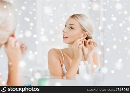beauty, jewelry and people concept - smiling young woman trying on diamond earring and looking to mirror at home bathroom over snow