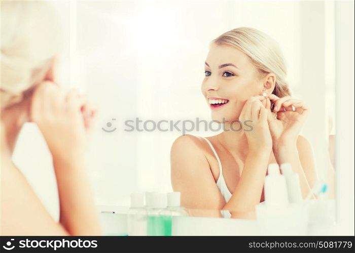 beauty, jewelry and people concept - smiling young woman trying on diamond earring and looking to mirror at home bathroom. woman trying on earring looking at bathroom mirror