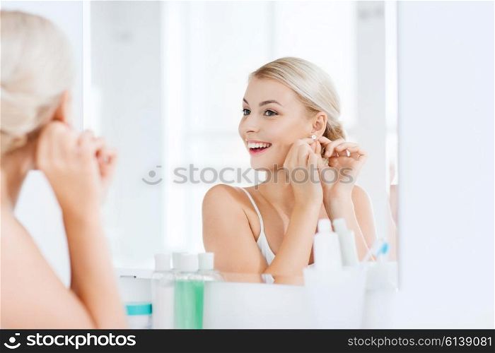 beauty, jewelry and people concept - smiling young woman trying on diamond earring and looking to mirror at home bathroom