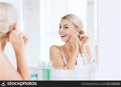 beauty, jewelry and people concept - smiling young woman trying on diamond earring and looking to mirror at home bathroom
