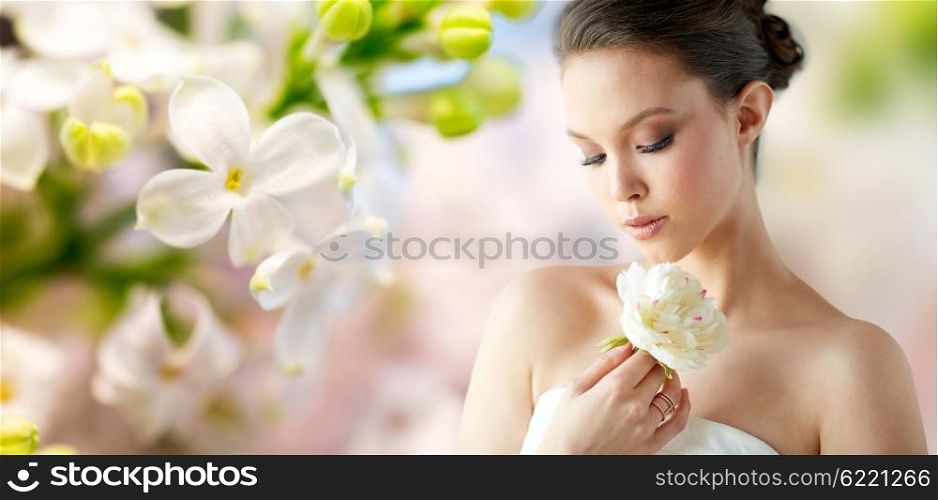 beauty, jewelry, people and luxury concept - beautiful asian woman or bride in white dress with peony flower and golden ring over natural spring lilac blossom background