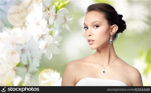 beauty, jewelry, wedding accessories, people and luxury concept - beautiful asian woman or bride with earring and pendant over natural spring cherry blossom background