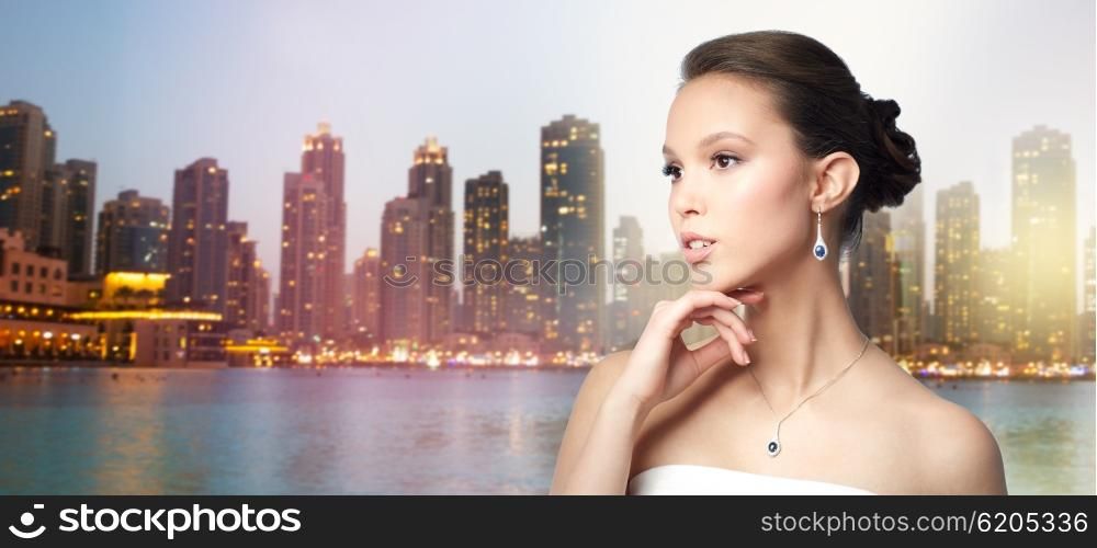 beauty, jewelry, wedding accessories, people and luxury concept - beautiful asian woman or bride with earring and pendant over evening dubai city lights and skyscrapers background
