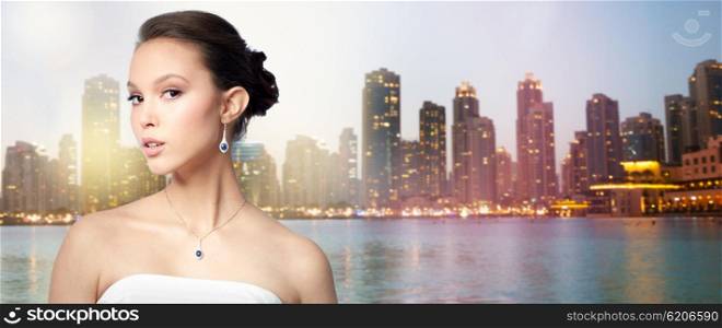 beauty, jewelry, wedding accessories, people and luxury concept - beautiful asian woman or bride with earring and pendant over evening dubai city lights and skyscrapers background