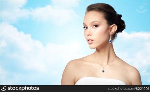 beauty, jewelry, wedding accessories, people and luxury concept - beautiful asian woman or bride with earring and pendant over blue sky and clouds background