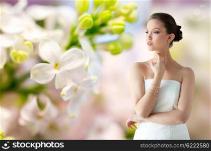 beauty, jewelry, wedding accessories, people and luxury concept - beautiful asian woman in white dress or bride with golden bracelet over natural spring lilac blossom background