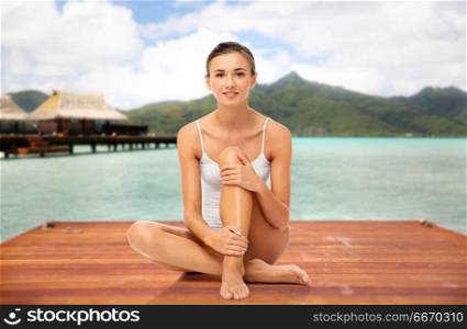 beauty, luxury and bodycare concept - beautiful woman touching her smooth bare legs sitting on wooden pier over bungalow and island in french polynesia. beautiful woman touching her smooth bare legs. beautiful woman touching her smooth bare legs