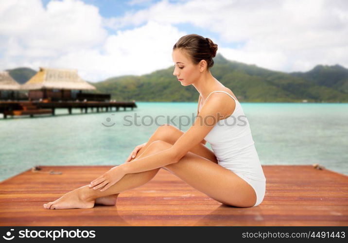 beauty, luxury and bodycare concept - beautiful woman touching her smooth leg skin sitting on wooden pier over bungalow and island in french polynesia. beautiful woman touching her smooth leg skin. beautiful woman touching her smooth leg skin