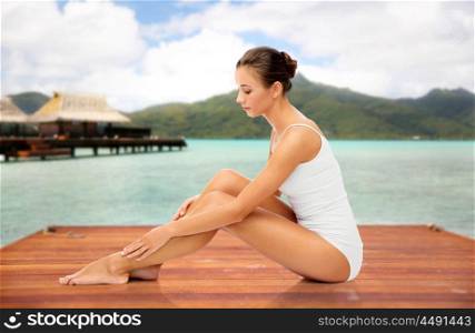 beauty, luxury and bodycare concept - beautiful woman touching her smooth leg skin sitting on wooden pier over bungalow and island in french polynesia. beautiful woman touching her smooth leg skin. beautiful woman touching her smooth leg skin