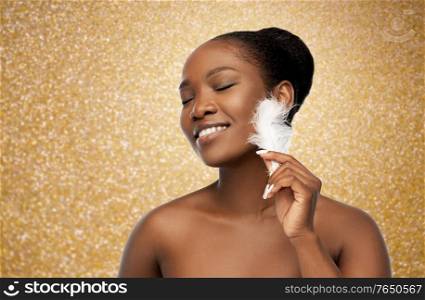 beauty, luxury and people concept - portrait of happy smiling young african american woman with bare shoulders and feather over golden glitter on background. happy african american woman with feather