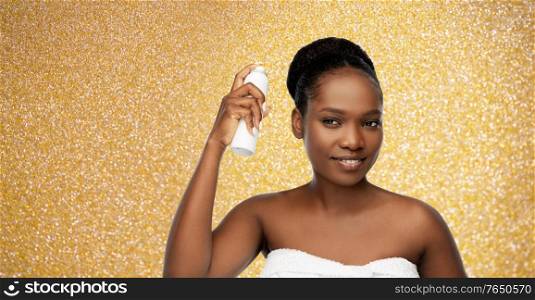 beauty, luxury and people concept - portrait of happy smiling young african american woman with bare shoulders using hairspray over golden glitter background. young african woman with hairspray