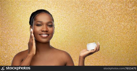 beauty, luxury and people concept - portrait of happy smiling young african american woman with bare shoulders applying moisturizer to her face over golden glitter background. smiling african american woman with moisturizer