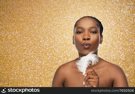 beauty, luxury and people concept - portrait of young african american woman with bare shoulders blowing to feather over golden glitter on background. african american woman with feather