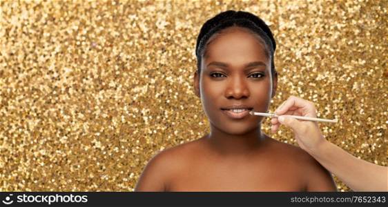 beauty, luxury cosmetics and people concept - close up of face of beautiful african american woman and hand with make up brush applying lipstick or gloss to lips over golden glitter background. face of african woman and hand with make up brush