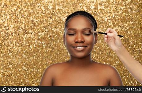 beauty, luxury cosmetics and people concept - portrait pf of beautiful smiling young african american woman and hand with make up brush applying eye shadow over golden glitter background. face of african woman and hand with make up brush