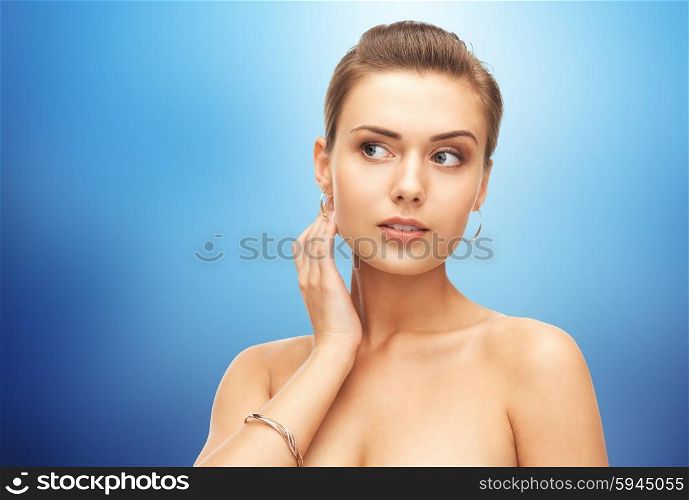 beauty, luxury, people, holidays and jewelry concept - beautiful woman wearing gold earrings and bracelet over blue background