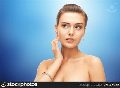 beauty, luxury, people, holidays and jewelry concept - beautiful woman wearing gold earrings and bracelet over blue background