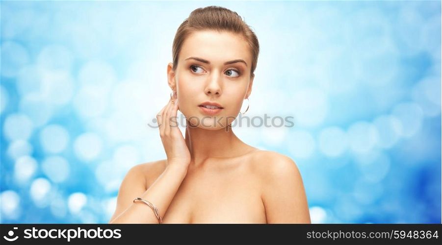 beauty, luxury, people, holidays and jewelry concept - beautiful woman wearing gold earrings and bracelet over blue lights background