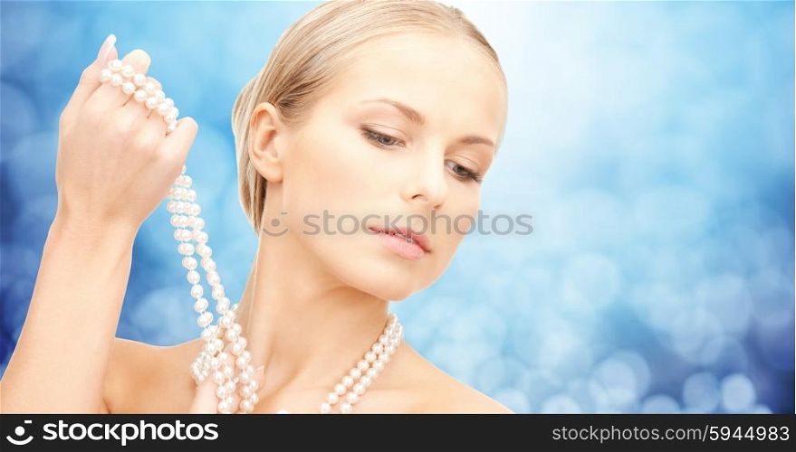 beauty, luxury, people, holidays and jewelry concept - beautiful woman with sea pearl necklace or beads in hand over blue lights background