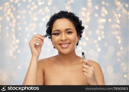 beauty, make up and cosmetics concept - beautiful young african american woman applying mascara over festive lights on grey background. beautiful african american woman applying mascara