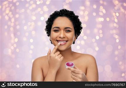 beauty, make up and cosmetics concept - beautiful young african american woman applying lip gloss with finger over festive lights on lilac background. african american woman applying lip gloss