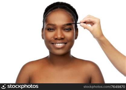 beauty, make up and cosmetics concept - portrait of smiling young african american woman and hand with mascara brush applying eyebrow shadows over white background. face of african woman and hand with mascara brush