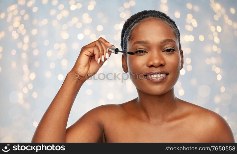 beauty, make up and cosmetics concept - smiling beautiful young african american woman applying mascara over festive lights background. beautiful african american woman applying mascara