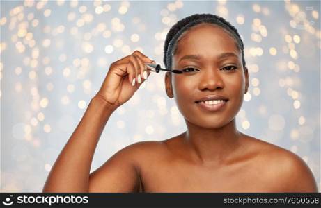 beauty, make up and cosmetics concept - smiling beautiful young african american woman applying mascara over festive lights background. beautiful african american woman applying mascara