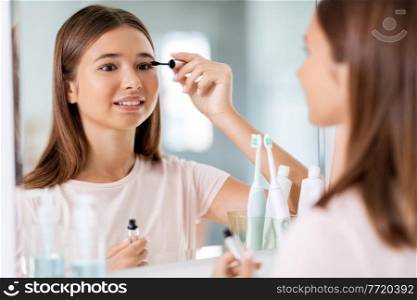beauty, make up and cosmetics concept - teenage girl applying eye makeup with mascara and looking to mirror at home bathroom. teenage girl applying mascara at bathroom