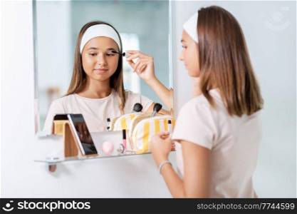 beauty, make up and cosmetics concept - teenage girl applying eye makeup with mascara and looking to mirror at home bathroom. teenage girl applying mascara at bathroom