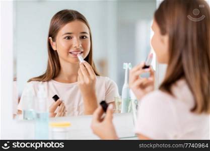 beauty, make up and cosmetics concept - teenage girl applying lipstick and looking to mirror at home bathroom. teenage girl applying lipstick at bathroom
