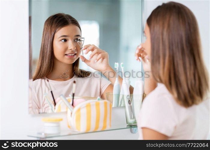 beauty, make up and cosmetics concept - teenage girl curling her lashes and looking to mirror at home bathroom. teenage girl curling her lashes at bathroom