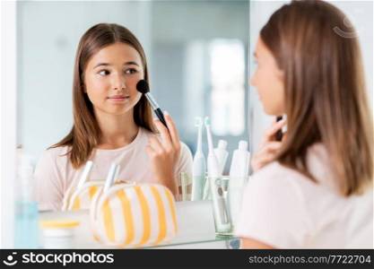 beauty, make up and cosmetics concept - teenage girl with brush applying blush to face and looking to mirror at home bathroom. teenage girl applying blush to face at bathroom