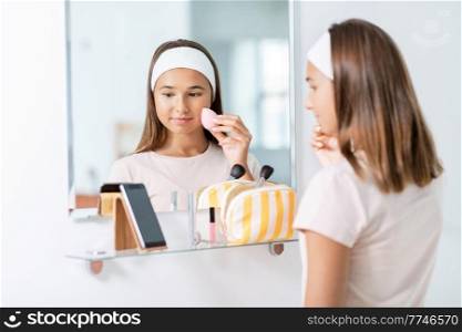 beauty, make up and cosmetics concept - teenage girl with sponge applying foundation to face and using smartphone in front of mirror at home bathroom. teenage girl with smartphone using make up sponge