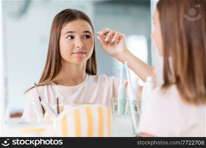 beauty, make up and cosmetics concept - teenage girl with tweezers tweezing her eyebrow and looking to mirror at home bathroom. teenage girl tweezing her eyebrow at bathroom