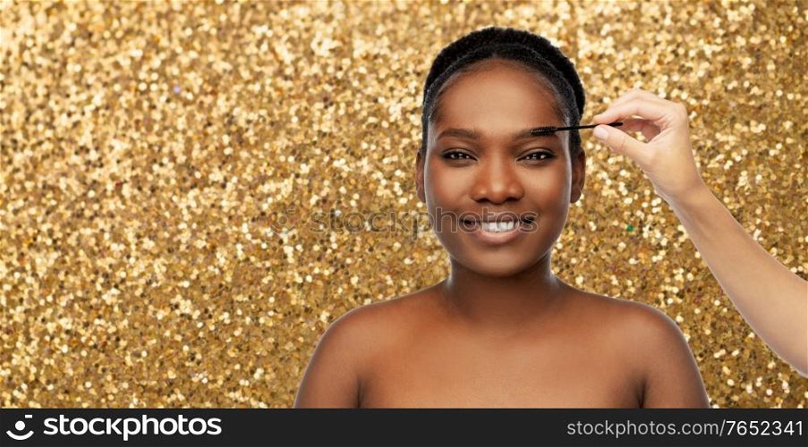 beauty, make up and luxury cosmetics concept - portrait of smiling young african american woman and hand with mascara brush applying eyebrow shadows over golden glitter background. face of african woman and hand with mascara brush
